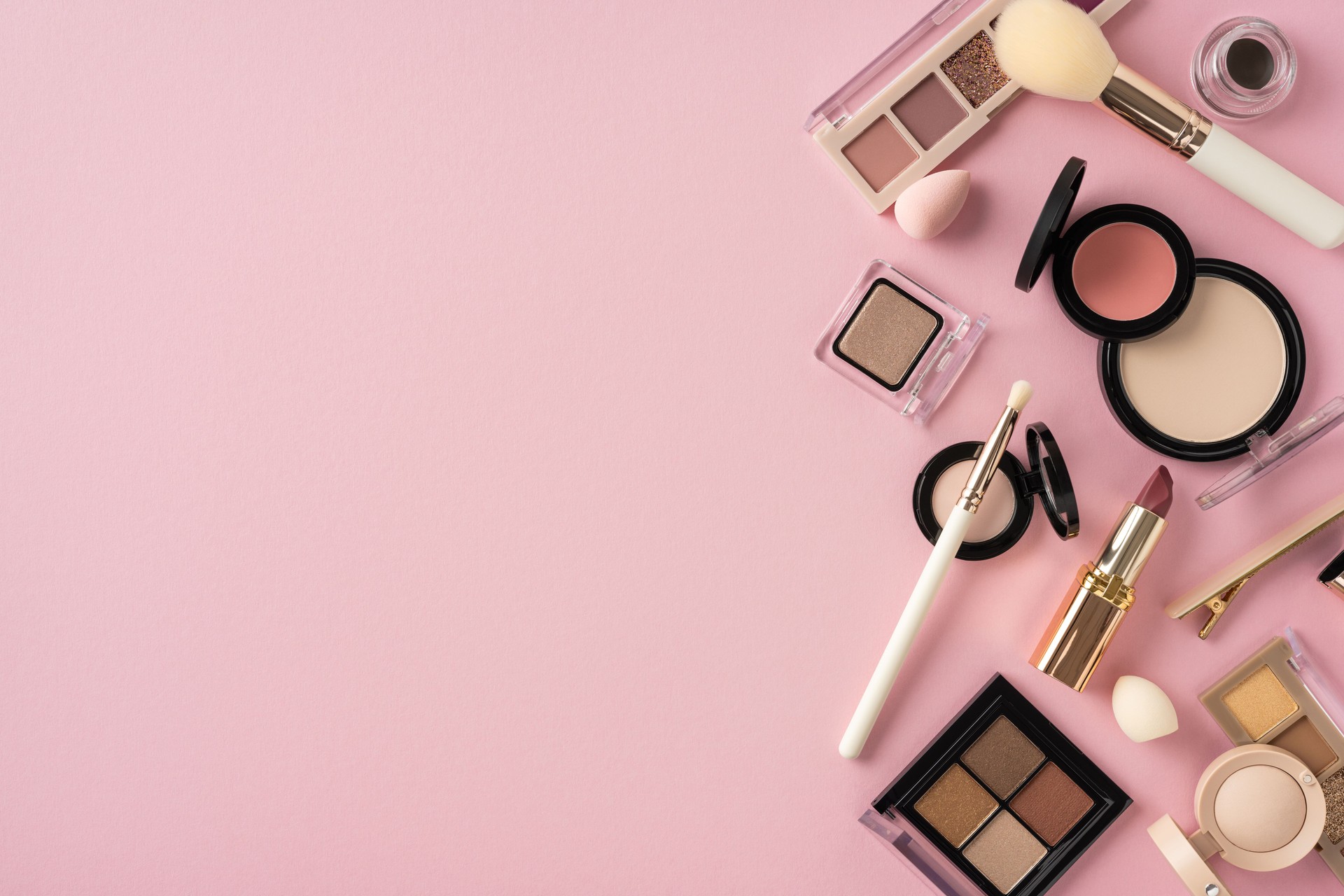 Assorted cosmetic items, including brushes, eyeshadows, and lipsticks, laid on pink backdrop